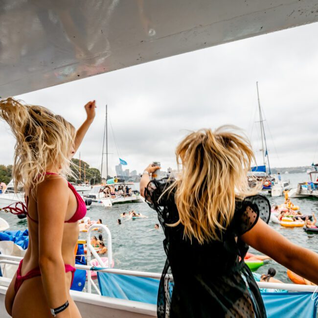 Two women in swimsuits dance energetically on a boat, their hair flying. Surrounding them are other boats and people swimming or lounging on inflatables in the water. In the background, trees line the shore beneath a cloudy sky. Experience this with Luxury Yacht Rentals Sydney at The Yacht Social Club.