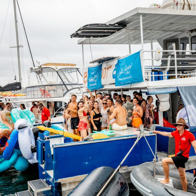 A lively group of people are gathered on and around a yacht, enjoying a festive atmosphere. The boat is decked with blue banners reading "The Yacht Social Club Event Boat Charters" and various inflatable pool toys are visible. The sky is cloudy, but the mood is jubilant and energetic.