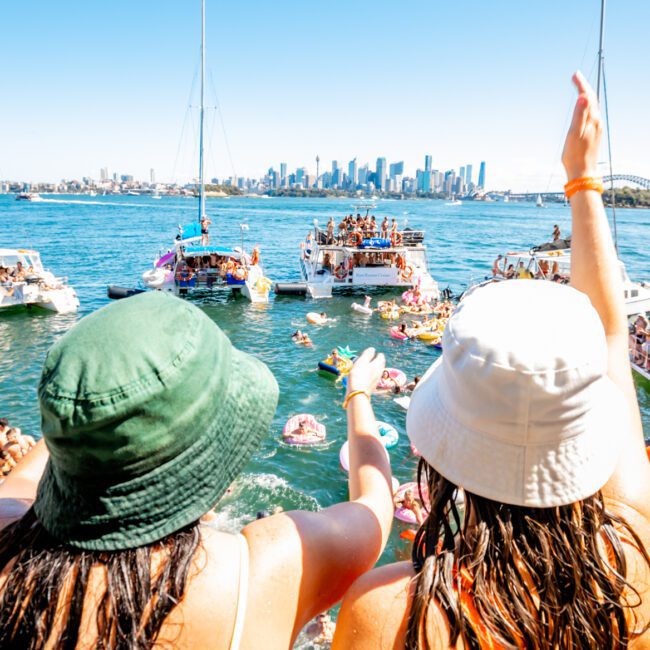 People are gathered on boats and floating in the water, enjoying a sunny day. Two individuals in the foreground, wearing hats, are waving towards other revelers. The skyline of a city is visible in the background under a clear blue sky, showcasing Luxury Yacht Rentals Sydney by The Yacht Social Club.