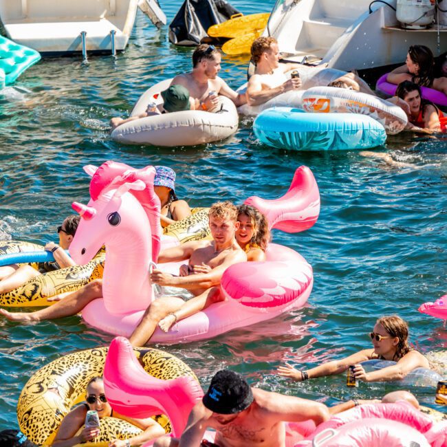 A lively group of people enjoy a sunny day on the water, lounging on inflatable pool floats, including an eye-catching pink unicorn. They are smiling, chatting, and holding drinks near boats from The Yacht Social Club Event Boat Charters, with others in colorful floats and swim rings surrounding them.