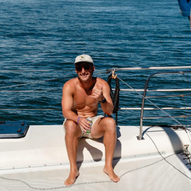 A man wearing a cap and sunglasses sits shirtless on a boat, smiling and holding a drink. The Yacht Social Club Sydney Boat Hire offers premium rentals for unforgettable moments like these. In the background, a cityscape and another boat grace the partly cloudy sky.
