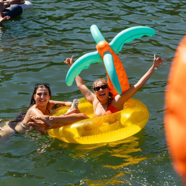 Two women are floating in a yellow inflatable ring with palm tree decorations on a lake, smiling. One makes a peace sign while the other raises her arm. Both wear sunglasses and swimwear. Other people on inflatables are visible in the background, capturing the fun vibe of The Yacht Social Club Boat Parties Sydney.