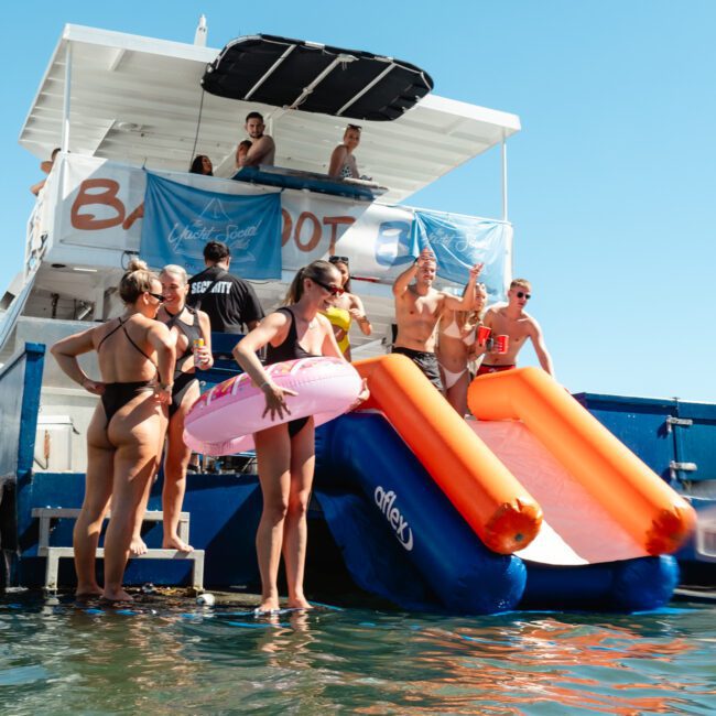 A group of people enjoys a sunny day on the water, gathered around a docked boat with a blue and orange slide. Some are in swimsuits, with one person holding a pink inflatable ring. Others are on the upper deck of the boat, under a canopy. The water is calm.