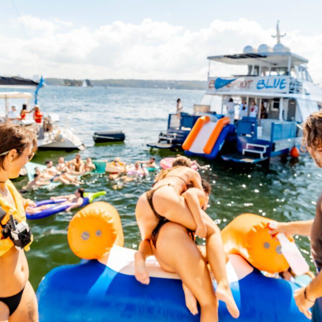 A lively scene unfolds on the water as a group of people enjoy a sunny day. Several individuals swim nearby while others relax on inflatable toys. Two boats are anchored, with one hosting a slide. The clear sky with scattered clouds adds to the charm of the day at The Yacht Social Club - Sydney Harbour Boat Hire.