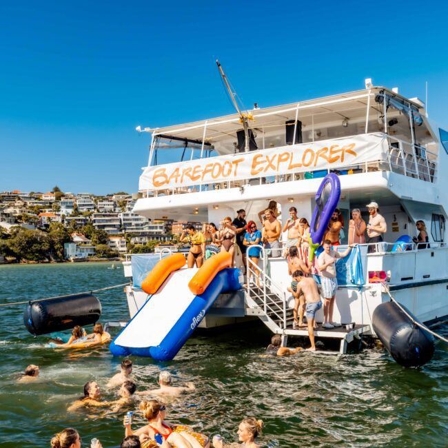 A lively scene of people enjoying a party on a large boat named "Barefoot Explorer." The Yacht Social Club Sydney Boat Hire offers slides, with some guests swimming and others relaxing on inflatables. Coastal houses and greenery are visible in the background under a sunny sky.