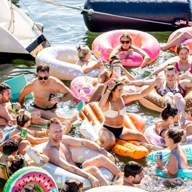 A group of people enjoying a sunny day in the water, sitting on colorful inflatable pool floats including a donut, watermelon slice, and pizza slice. They are smiling, chatting, and holding drinks, with a boat from Sydney Harbour Boat Hire The Yacht Social Club partially visible in the background.