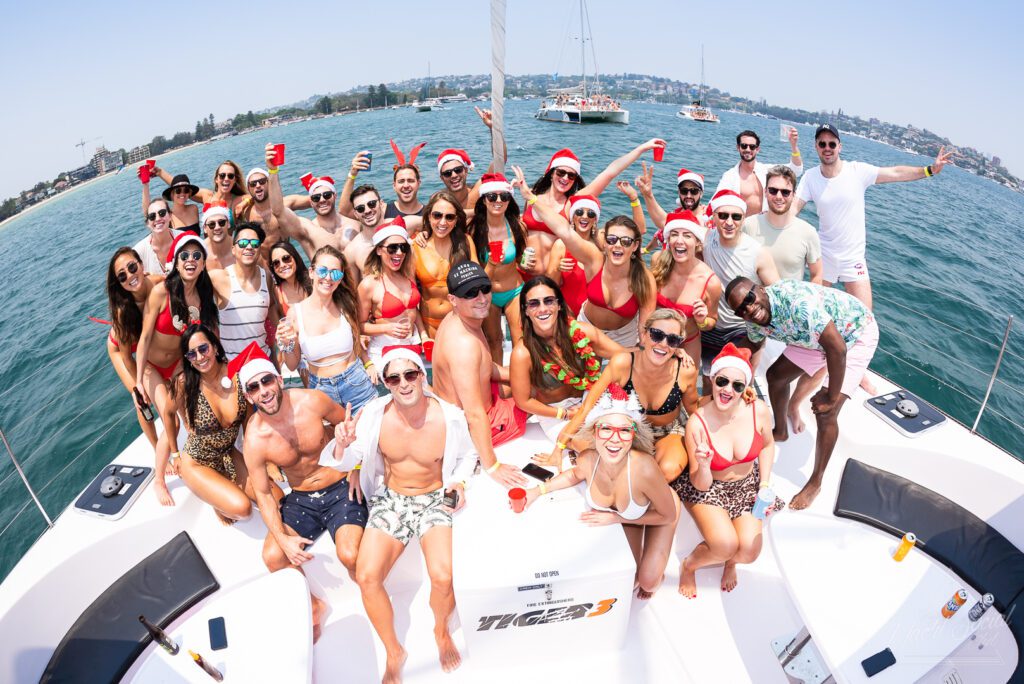 A large group of people, many wearing Santa hats and festive attire, are gathered on a boat. They're smiling, raising drinks, and posing for the camera. The Yacht Social Club Sydney Boat Hire sets the scene on a sunny day with other boats and a distant shoreline visible under a clear sky.