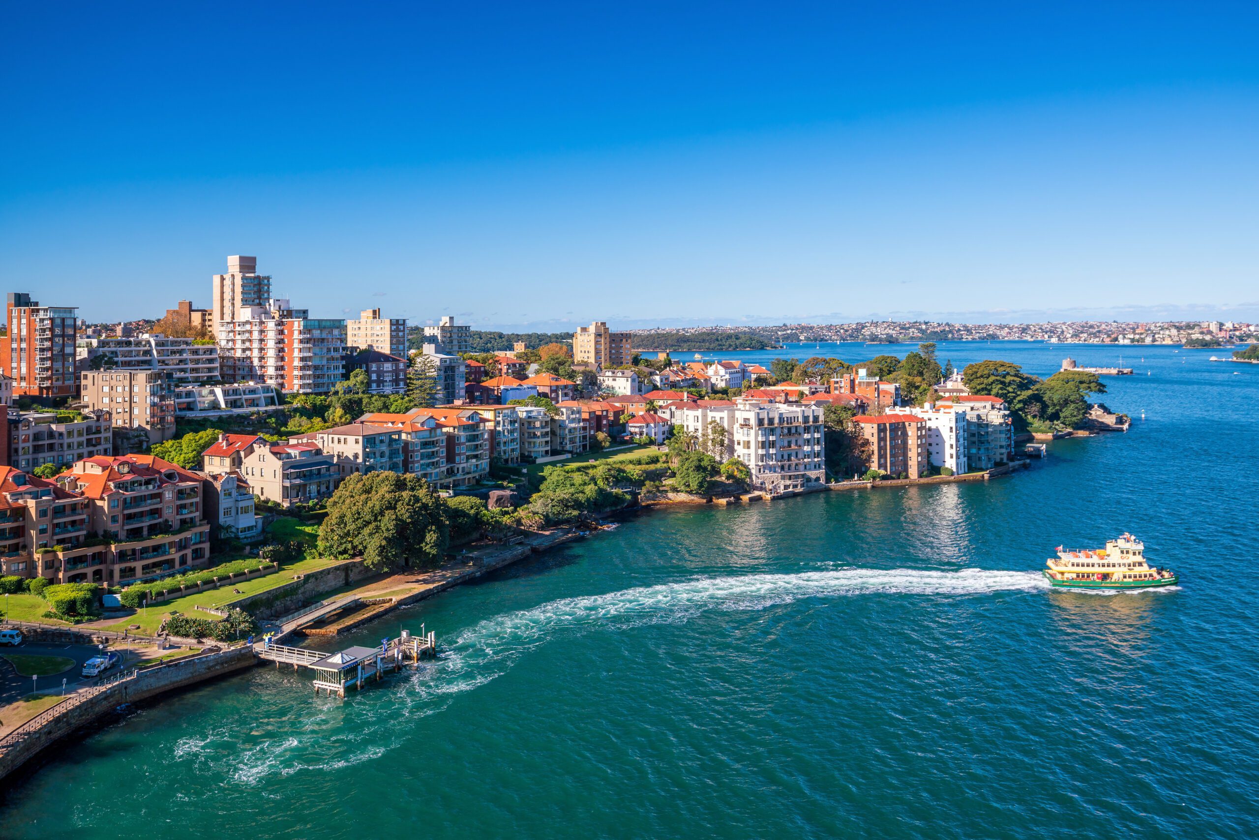 A scenic view of a harbor with clear blue water and a boat from The Yacht Social Club leaving a wake behind it. Residential buildings with red-tiled roofs and greenery line the waterfront. The background features more buildings, green spaces, and a vast blue sky.