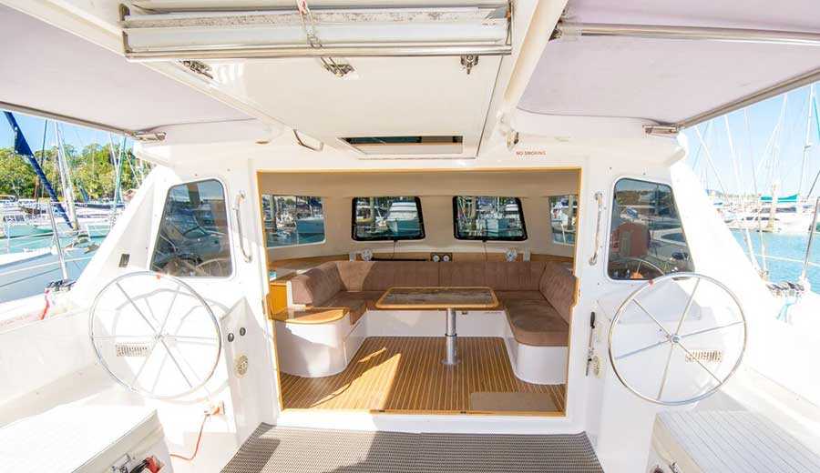 Interior view of a boat with two steering wheels, a table, and a seating area covered in beige cushions. The boat is at a marina, with windows providing a view of the surrounding water and docked boats. Light floods the clean, polished space—perfect for Luxury Yacht Rentals Sydney or The Yacht Social Club Sydney Boat Hire.