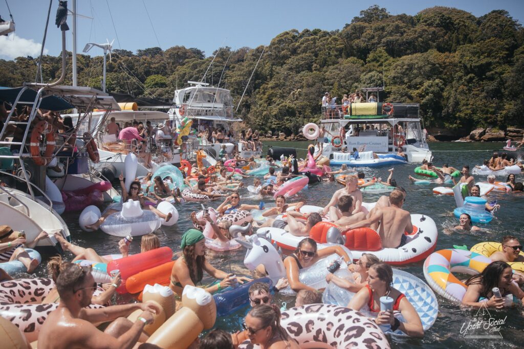 A large group of people are enjoying a boat party in the water, surrounded by various inflatables such as unicorns, doughnuts, and flamingos. Several boats from The Yacht Social Club Event Boat Charters are anchored nearby, and the shore is lined with lush trees under a clear sky.