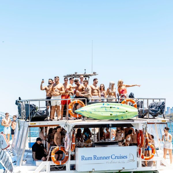 A large group of people in swimsuits stands on the upper deck of a boat named "Rum Runner Cruises," enjoying the sunny weather. Some passengers are posing and waving, with a city skyline and bridge visible in the background over the blue water. Experience unforgettable moments with The Yacht Social Club's Boat Parties Sydney.