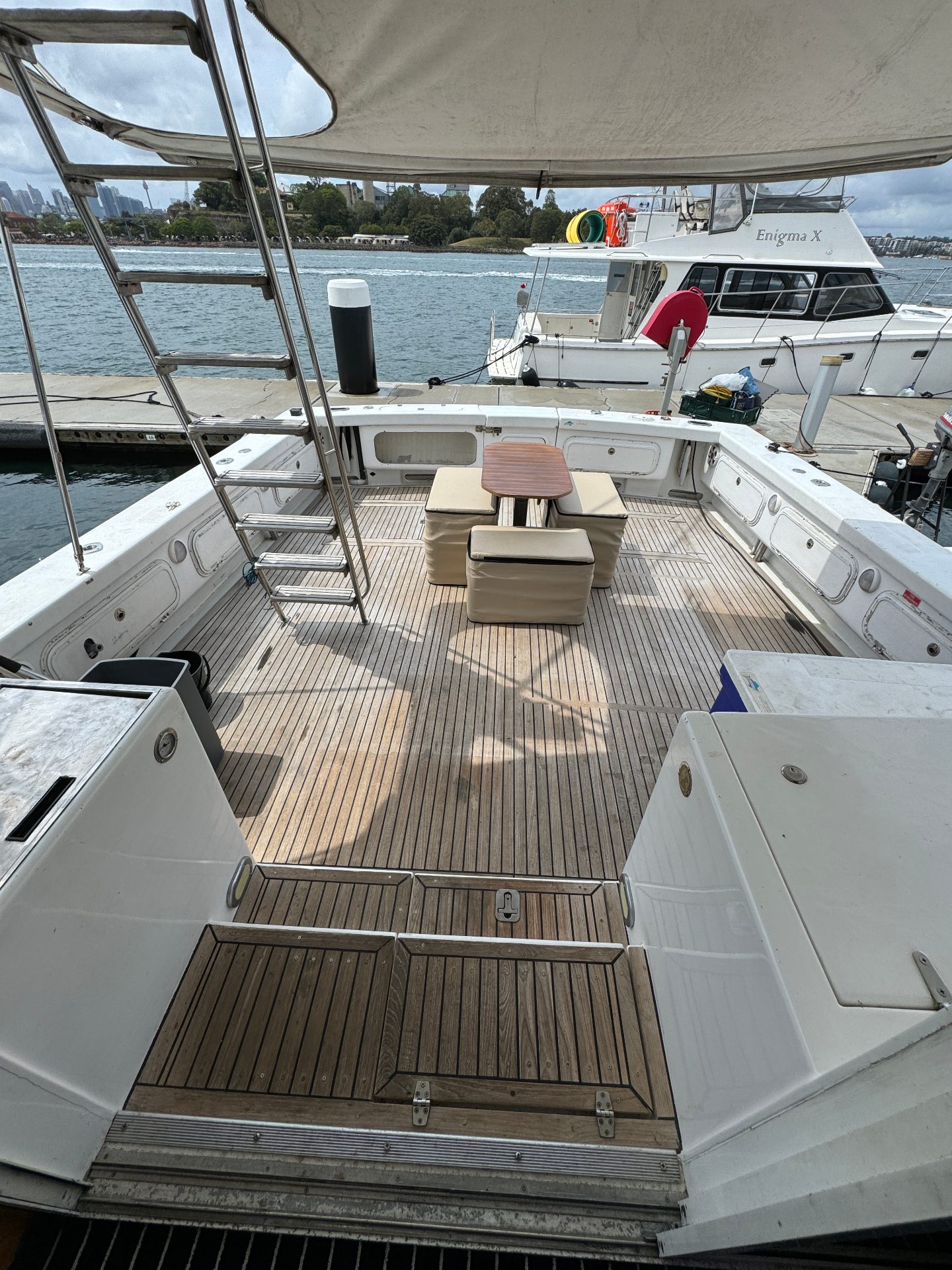 The Yackatoon features a spacious deck with wooden flooring, a small table, and cushioned seats. Docked at a marina, it offers views of boats and water, plus a metal ladder leading to the upper deck.