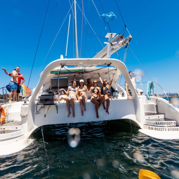 A group of people sitting and relaxing on the stern of a white catamaran named "Barefoot" with the registration "ON 862711 SYDNEY." The Yacht Social Club adds a touch of elegance to this sunny, scenic setting. Clear blue skies frame the boat, with another vessel visible in the background.