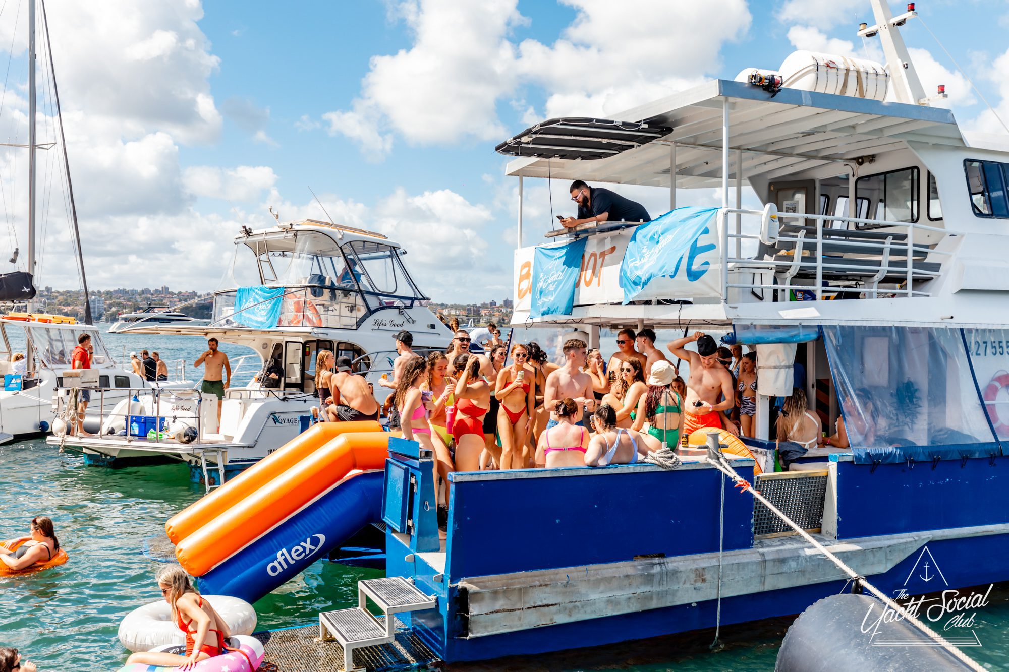 A lively boat party hosted by The Yacht Social Club features groups of people on a docked two-level boat. Many wear swimwear, socializing and enjoying the sun. An inflatable orange and blue slide leads from the boat into the water as other boats float nearby under a partly cloudy sky.