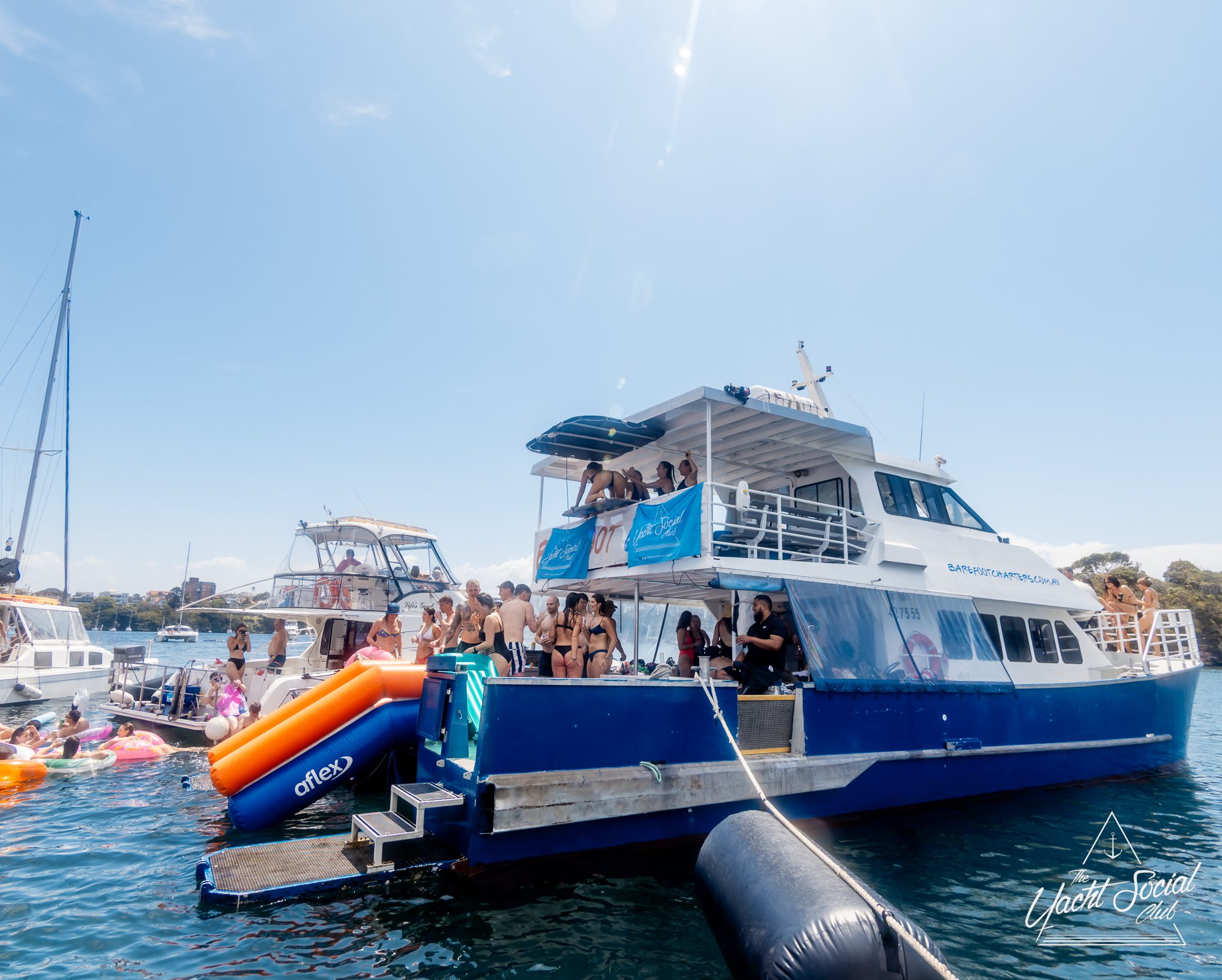 A large blue and white multi-deck yacht from The Yacht Social Club Sydney Boat Hire is anchored on the water, bustling with people celebrating. Some enjoy the upper deck, while others relax on inflatable floats nearby. A clear blue sky and other boats complete the vibrant scene.