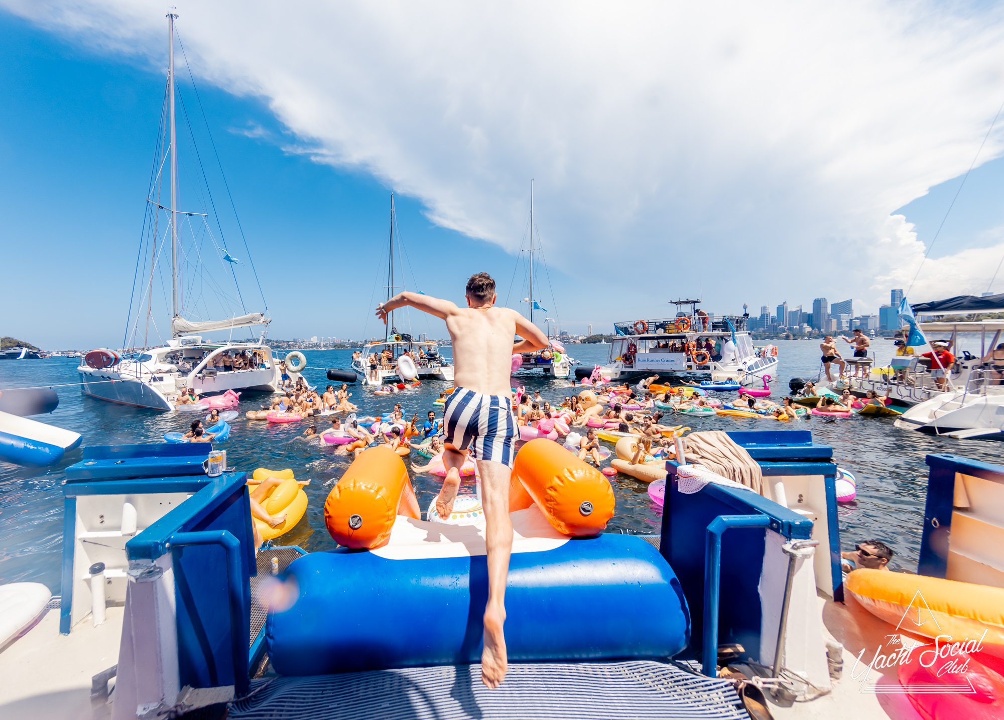 A person in striped swim trunks jumps from a boat into a lively and colorful gathering of people on inflatable floats in the water, with various boats anchored nearby. The backdrop features a city skyline and a dramatic sky with fluffy clouds. It's an exciting scene at The Yacht Social Club Event Boat Charters.