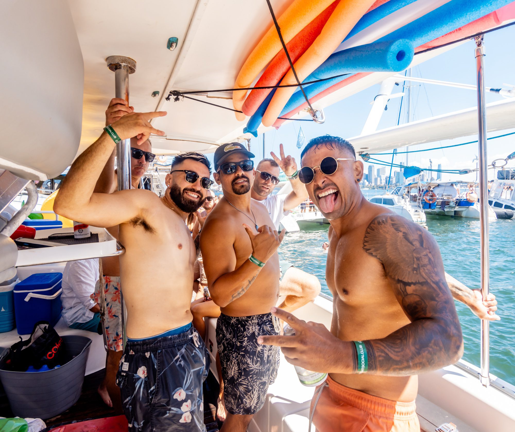 A group of shirtless men on a boat enjoy a sunny day. They are smiling, posing for the camera, and surrounded by colorful pool noodles. Wearing sunglasses and casual swimwear, they revel in the relaxed and celebratory vibe with sailboats in the background, courtesy of Sydney Harbour Boat Hire The Yacht Social Club.