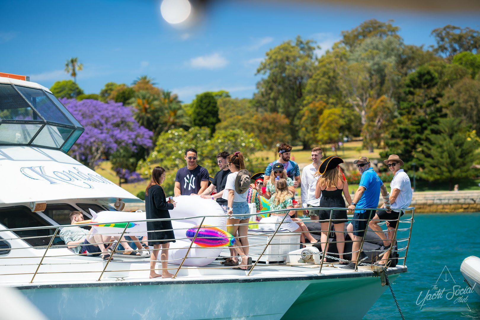 A group of people enjoying a sunny day on a yacht from The Yacht Social Club Sydney Boat Hire. They are gathered on the deck, some standing and others sitting, engaging in conversation. The yacht is on calm water with scenic trees in the background. Inflatable pool toys can be seen on the deck.