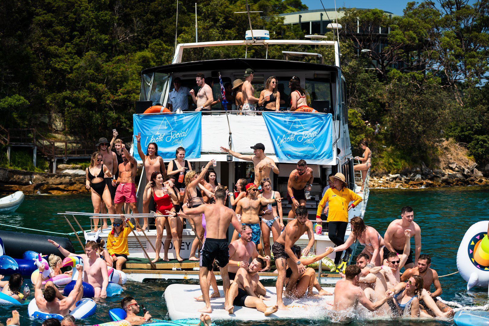 A large group of people in swimwear are enjoying a lively boat party. Some are dancing on the deck, while others float on inflatables. Two blue banners with text are displayed on the railing. Dense greenery is visible in the background. Hosted by The Yacht Social Club Sydney Boat Hire, it’s a perfect event for fun and relaxation.