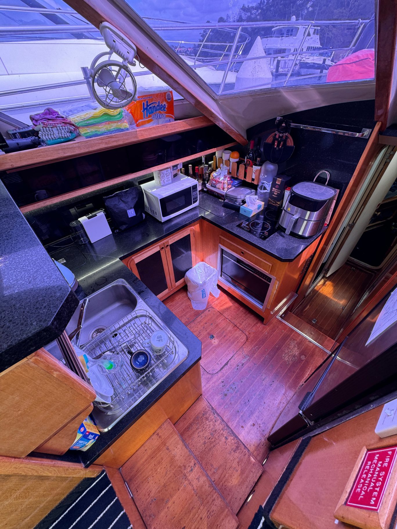 The Yackatoon features a compact boat kitchen with a sink, microwave, rice cooker, toaster, countertop kitchen items, a trash bin on the wooden floor, and a window displaying part of another boat outside.