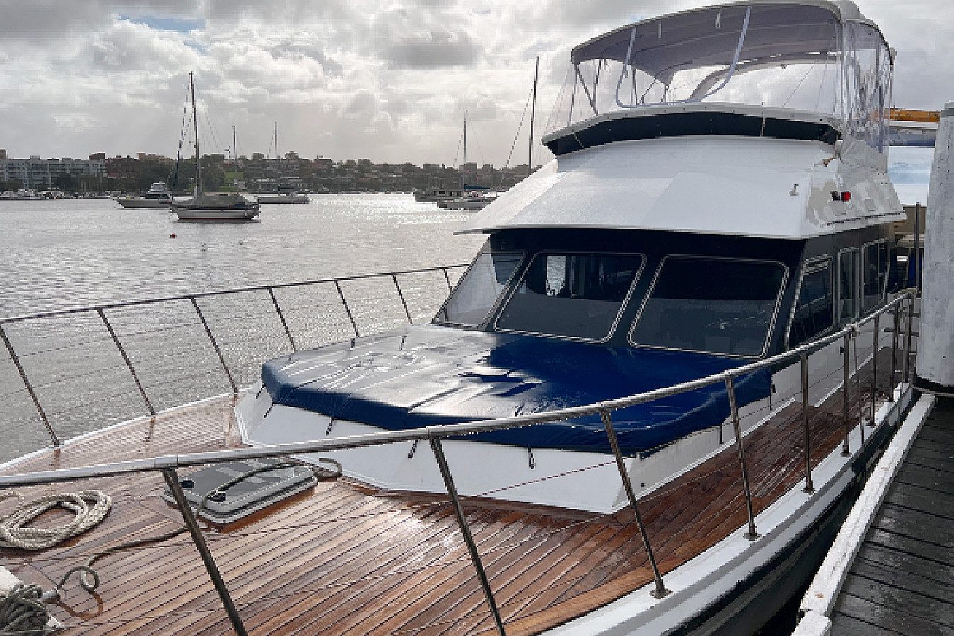 A docked motor yacht with a blue cover on the bow. The boat features a wooden deck and is tied to a dock. The water reflects the cloudy sky, and several sailboats are visible near a distant shore. Experience the charm of this scene with The Yacht Social Club Event Boat Charters in Sydney Harbour.