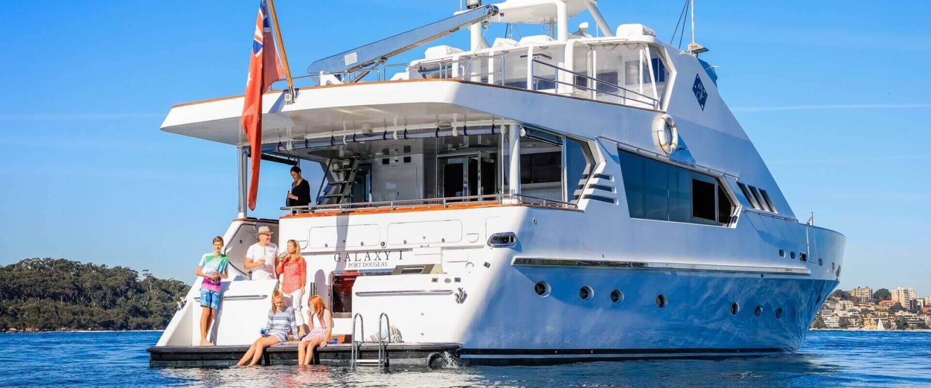 A large luxury yacht is anchored in clear water. Five people gather on the deck and swimming platform: some stand and chat, others sit with their feet in the water. The yacht, part of Boat Parties Sydney The Yacht Social Club, has multiple decks, large windows, and a flag flying at the rear. Trees and buildings are visible in the background.