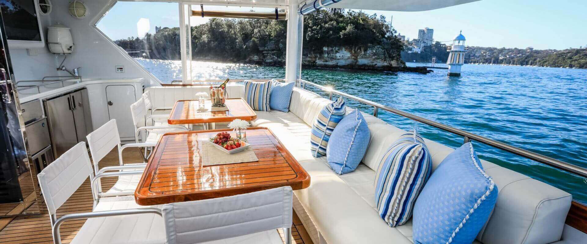 A luxurious yacht deck with a dining area featuring two polished wooden tables and white chairs. The built-in couch is adorned with blue and white striped pillows. Scenic view of calm waters, a small lighthouse, and distant shoreline in the background. Perfect for Boat Rental and Parties Sydney by The Yacht Social Club.