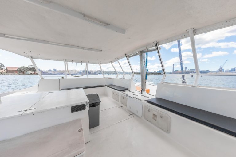 The interior of the boat features elegant white walls and seating, providing a wide view of the water and city skyline through large windows. The Yacht Social Club Event Boat Charters includes built-in benches, a central table, and a nearby trash can. The sky is partly cloudy.
