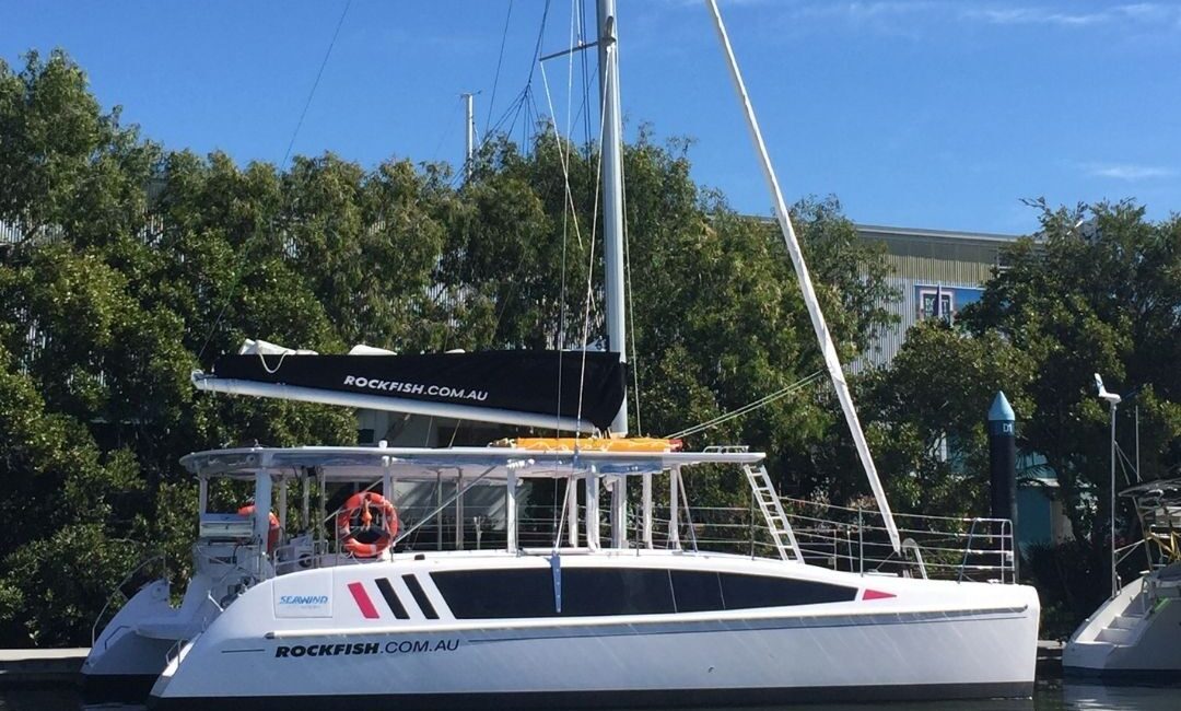 A white catamaran named "Rockfish.com.au" is docked at a marina on a sunny day. The vessel, often featured in Sydney Harbour Boat Hire The Yacht Social Club events, has a black sail and red accents on its hull. Trees and a marina building can be seen in the background.