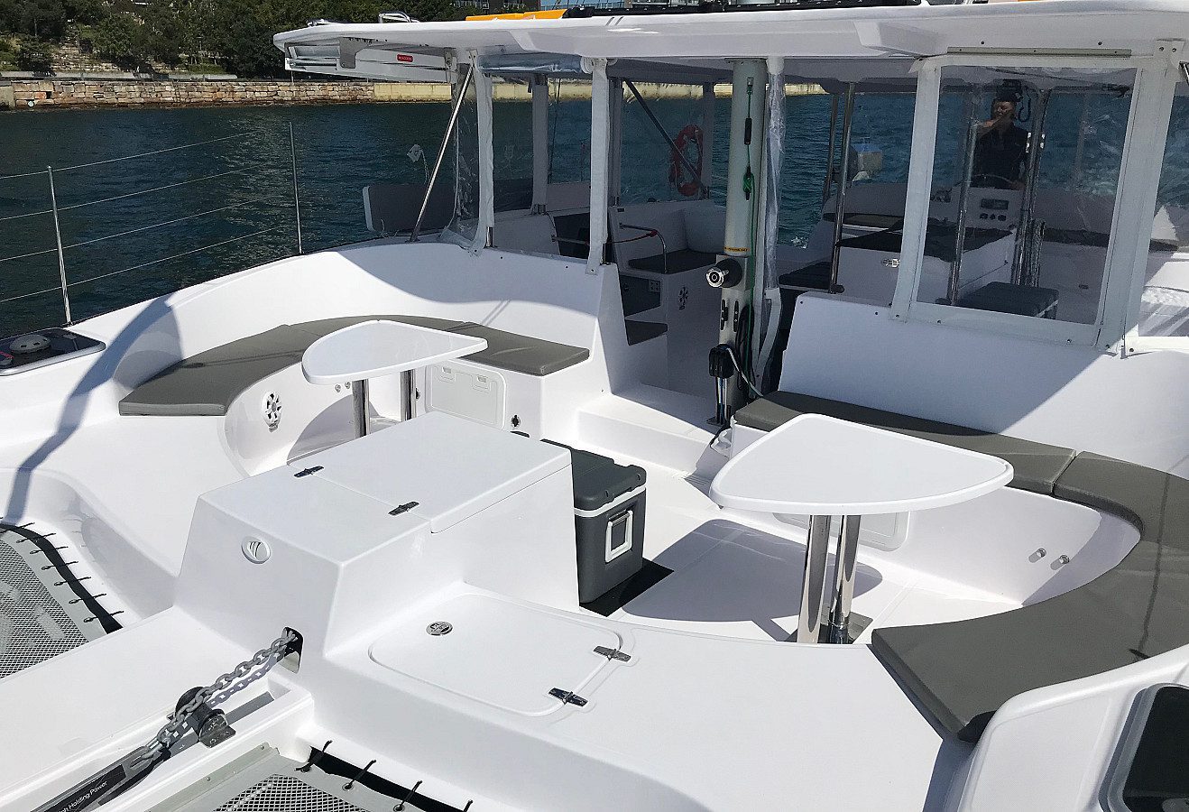 The image showcases the chic outdoor seating area of a catamaran, ideal for indulging in Sydney's famous boat parties with Luxury Yacht Rentals Sydney. The white, clean deck features cushioned benches and two small white tables. There are also storage compartments and other nautical equipment visible against a backdrop of water and rocky shoreline.