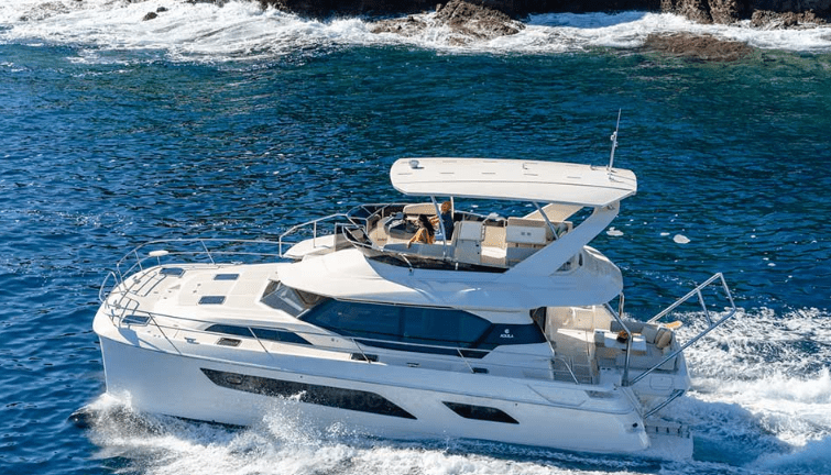 The Allure, a white luxury yacht, glides through clear blue ocean waters near a rocky shore, leaving a trail of white foam behind under the sun.