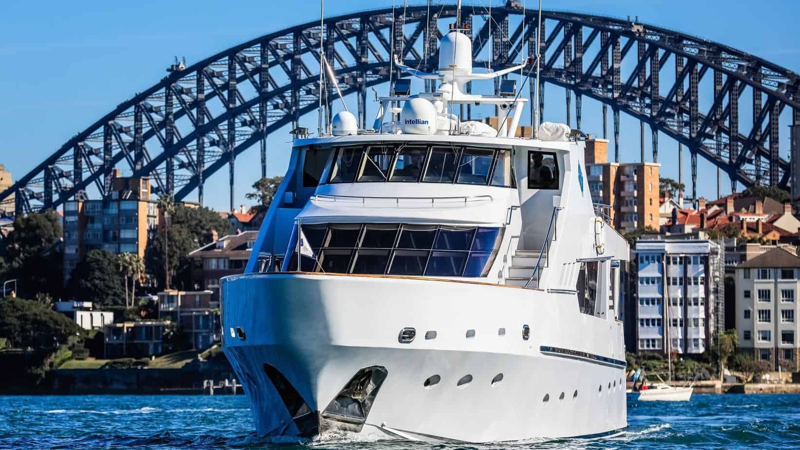 A large white yacht cruises through the water with a cityscape and iconic steel arch bridge in the background. The sunny day highlights the blue sky and calm waters. Residential buildings line the shore, offering a picturesque view perfect for The Yacht Social Club Event Boat Charters in Sydney.