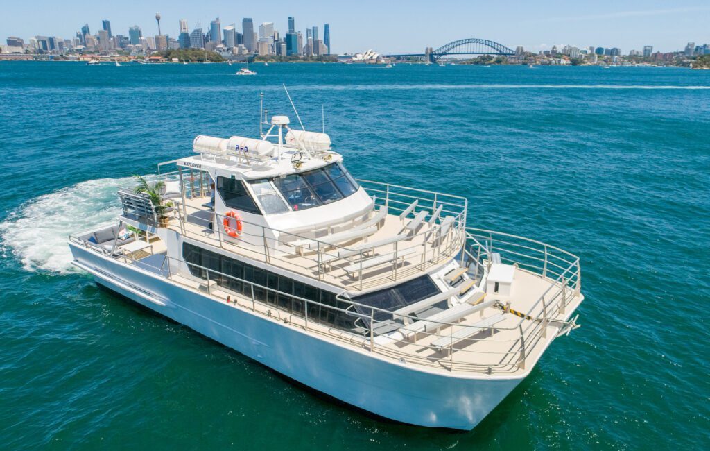 A white yacht from Sydney Harbour Boat Hire The Yacht Social Club cruises on a blue ocean with a city skyline and a prominent bridge in the background. Lounge chairs adorn the deck as it moves at a moderate speed, leaving a visible wake behind.
