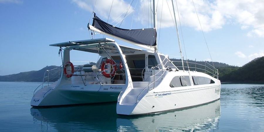 A sleek white catamaran with a black sail is anchored on calm, clear blue water. Two red lifebuoys are attached to its rear deck railing. The sky is partly cloudy, and lush green hills are visible in the background, perfect for Luxury Yacht Rentals Sydney or Boat Parties Sydney with The Yacht Social Club.