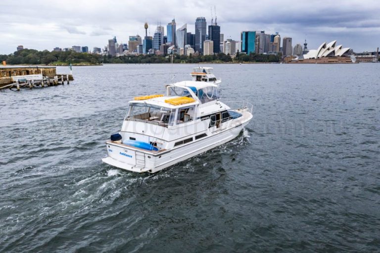 A white motorboat moves through the waters of a harbor with a city skyline in the background. Notable landmarks include the Sydney Opera House and tall skyscrapers. The sky is cloudy, and the water is slightly choppy. A pier extends from the left side of the image, perfect for parties hosted by The Yacht Social Club Sydney Boat Hire.