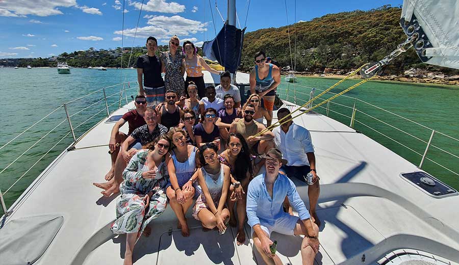 A group of people enjoying a sunny day with The Yacht Social Club Sydney Boat Hire. They are gathered on the deck, smiling and posing for a photo. The water and shoreline with greenery are visible in the background. It’s a clear day with blue skies and a few clouds, perfect for boat parties in Sydney.