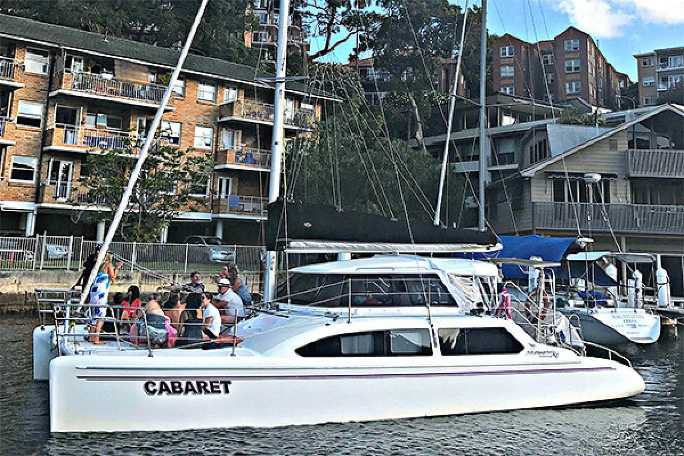 A group of people sit on a white boat named "Cabaret," docked alongside a marina with apartment buildings in the background. The boat, perfect for Boat Parties Sydney by The Yacht Social Club, features a sail and several windows. The water appears calm with surrounding greenery and multiple boats.