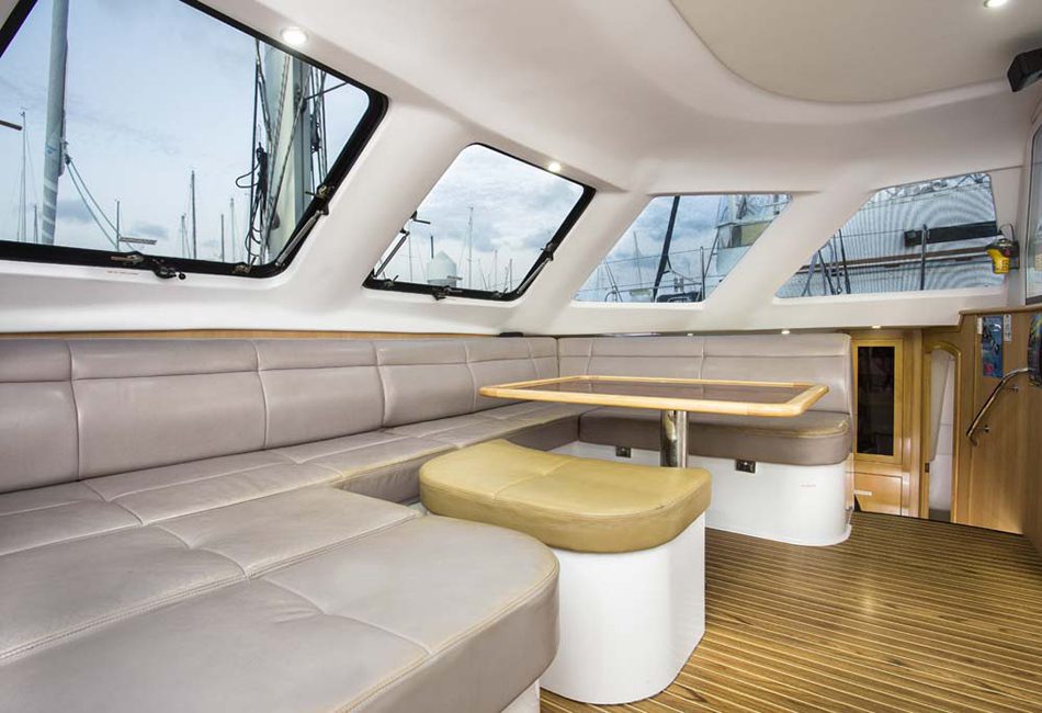 Interior of a modern boat with a seating area featuring light gray cushioned seats and a wooden table. Large windows provide ample natural light and views of the marina. The floor has a wood finish, and an adjacent door leads to the lower deck. Experience this at Luxury Yacht Rentals Sydney.
