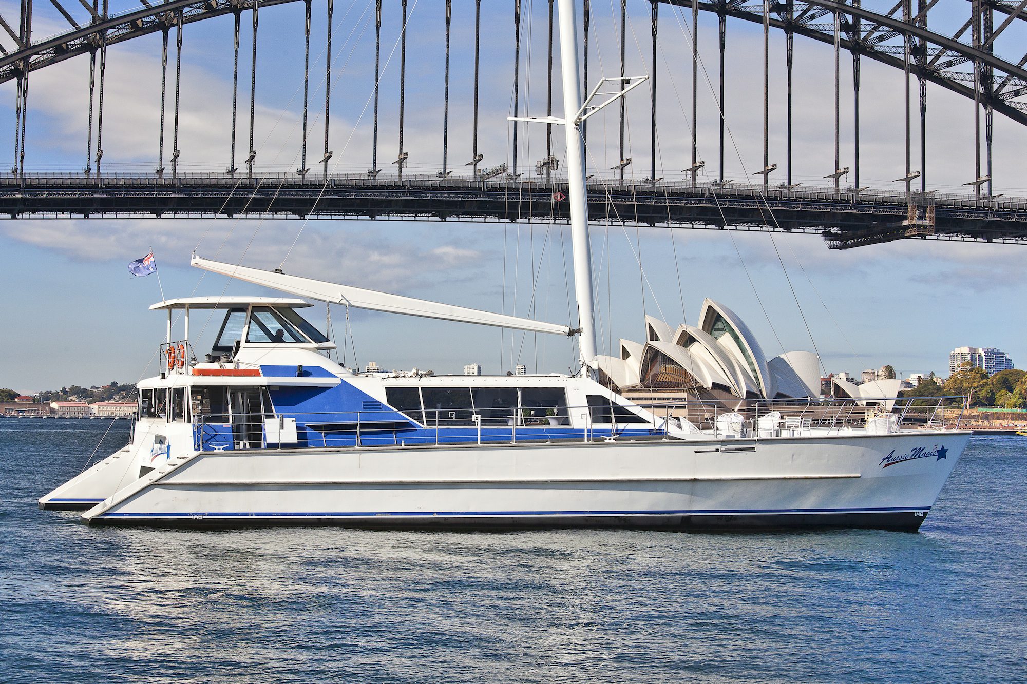 A white and blue catamaran sails on a sunny day, with the iconic Sydney Harbour Bridge and Sydney Opera House in the background. The boat, part of The Yacht Social Club, has a flag and several windows along its side. The water is calm and the sky partly cloudy.