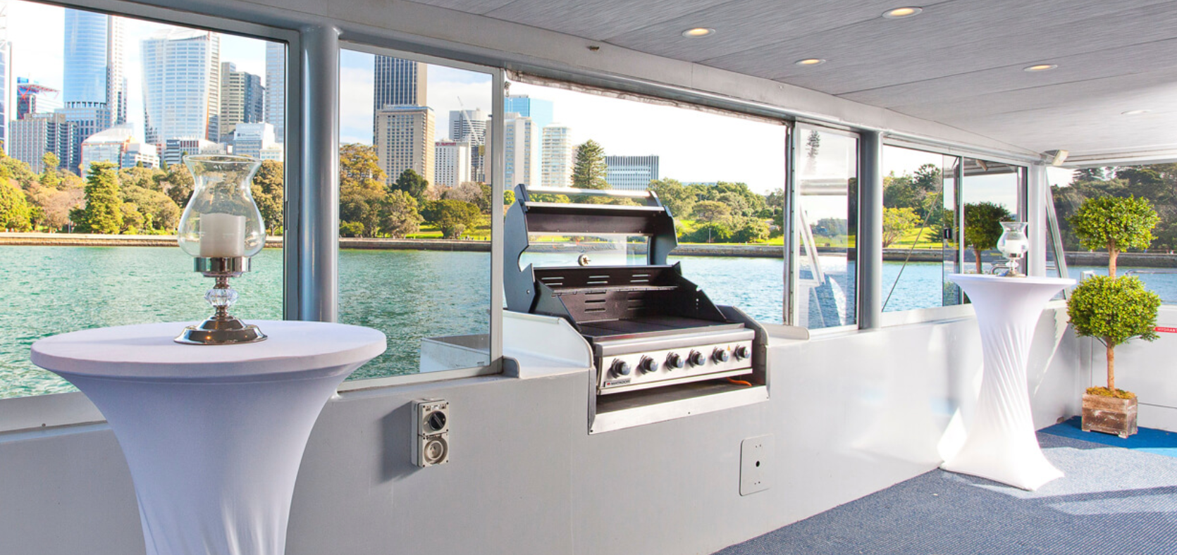 A modern indoor space with large windows overlooks a waterfront cityscape with trees and tall buildings. Inside, there is a barbecue grill and two tall cocktail tables covered in white cloth. Decorative lanterns and potted plants add an elegant touch, perfect for gatherings by The Yacht Social Club Sydney Boat Hire.