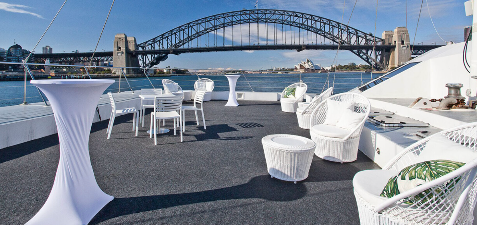 A rooftop deck on a boat with several white wicker chairs, tables, and high-top cocktail tables. The Yacht Social Club Sydney Boat Hire offers breathtaking views of the Sydney Harbour Bridge and the Sydney Opera House against a clear blue sky.