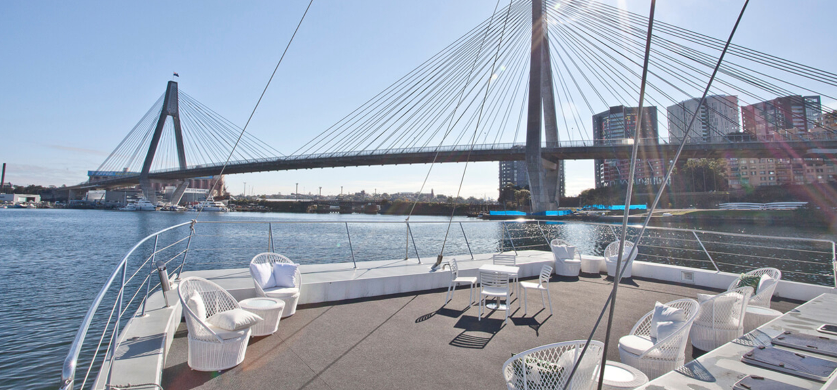 A spacious deck area on a boat features white wicker chairs and a table, overlooking calm waters with a large cable-stayed bridge and cityscape in the background under a clear blue sky. Experience the elegance of *Luxury Yacht Rentals Sydney* for an unforgettable day on the water.