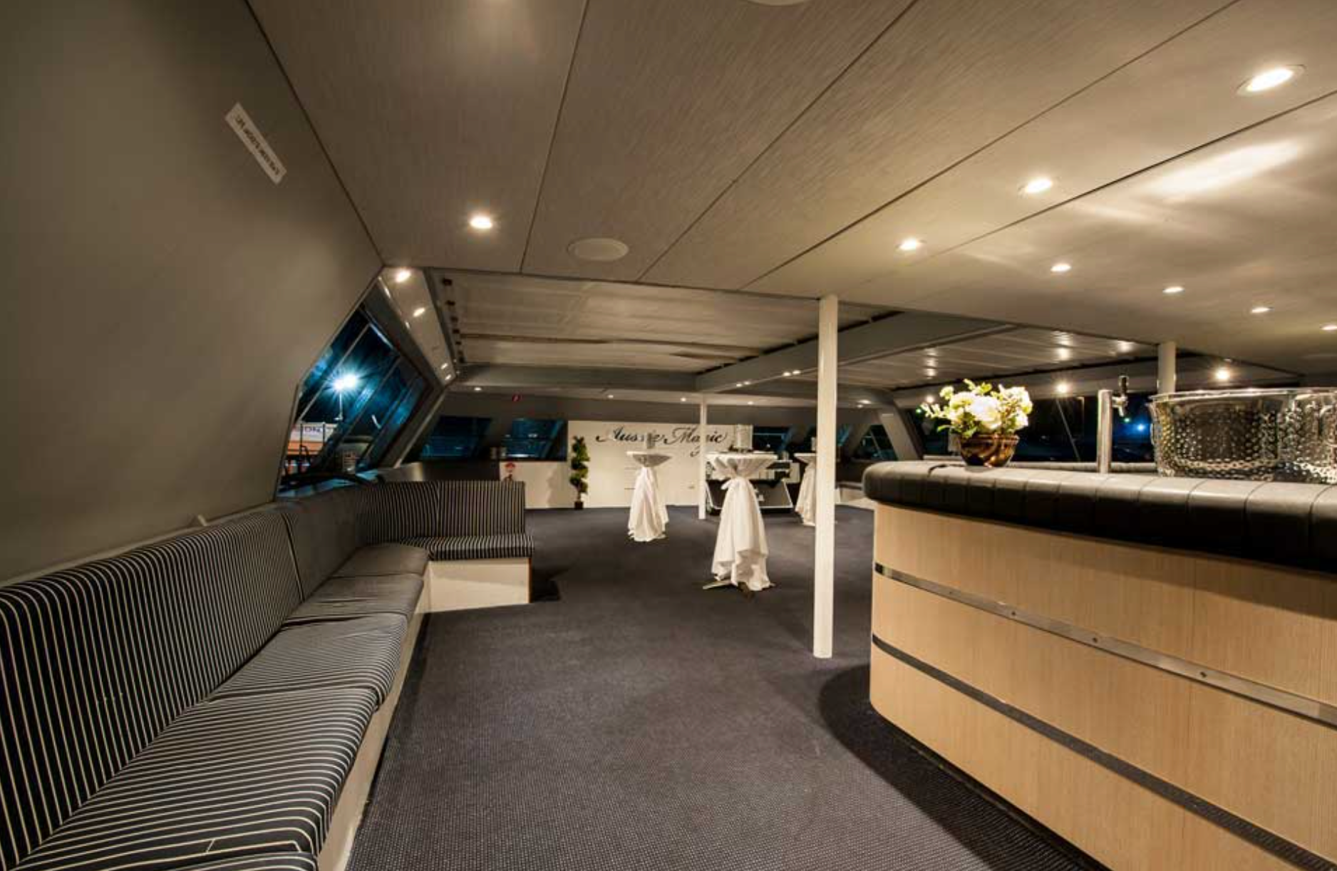 A modern boat interior with dim lighting features a sleek bar with a striped cushioned seat along the wall. There are small round tables with white tablecloths and flower arrangements. Large windows line the walls, and the ceiling is adorned with recessed lighting—perfect for Luxury Yacht Rentals Sydney.