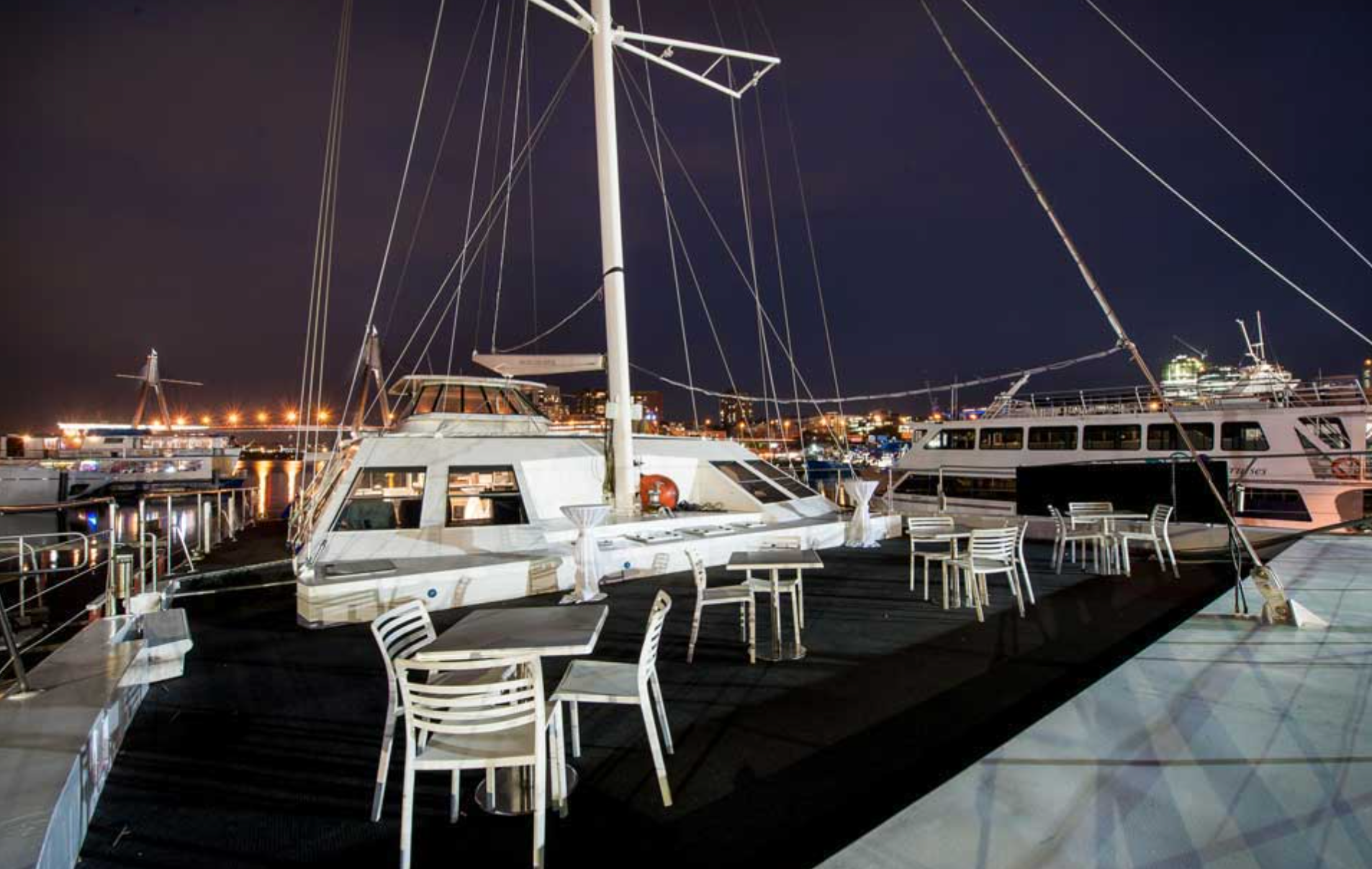 A well-lit marina at night features a large, white catamaran from Luxury Yacht Rentals Sydney docked next to several other boats. Empty white chairs and tables are arranged on the dock, suggesting an outdoor seating area. The background showcases city lights and buildings.
