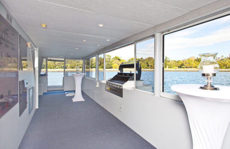 The Aussie Magic features a spacious boat deck with large windows, carpeted floors, white-covered cocktail tables, a decorative lamp, barbecue grill, and scenic views of water and greenery.