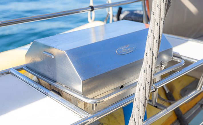 A rectangular, stainless steel grill is mounted on the railing of a luxury yacht from The Yacht Social Club Sydney Boat Hire. The shiny lid is closed, with ropes and the ocean visible in the background, suggesting the yacht is on water. Sunlight reflects off the grill in the bright setting.