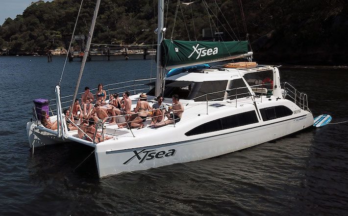 A group of people relaxes on the deck of a white Xsea catamaran sailboat anchored in a serene waterway. Trees and a small dock are visible in the background under a clear sky. Some are sunbathing while others sit under the green sail, enjoying The Yacht Social Club's Sydney Harbour Boat Hire experience.
