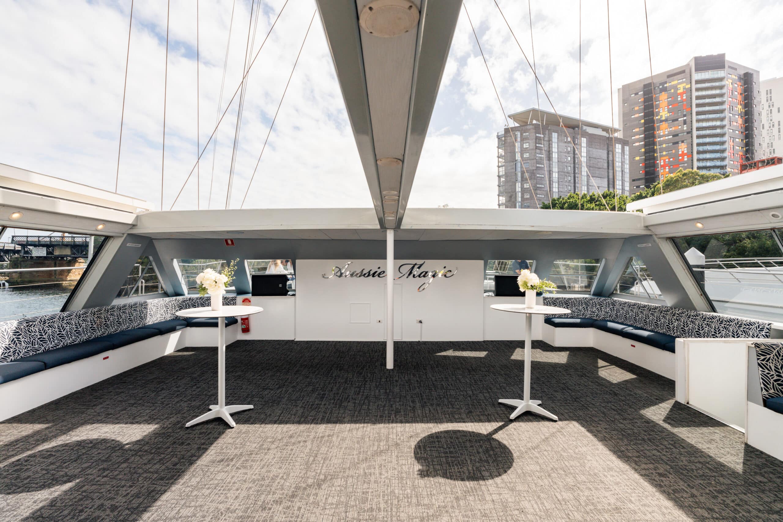 The Aussie Magic yacht features a spacious deck with grey carpet, blue cushioned side seating, two white cocktail tables adorned with flower vases, and city views set beneath a partly cloudy sky.