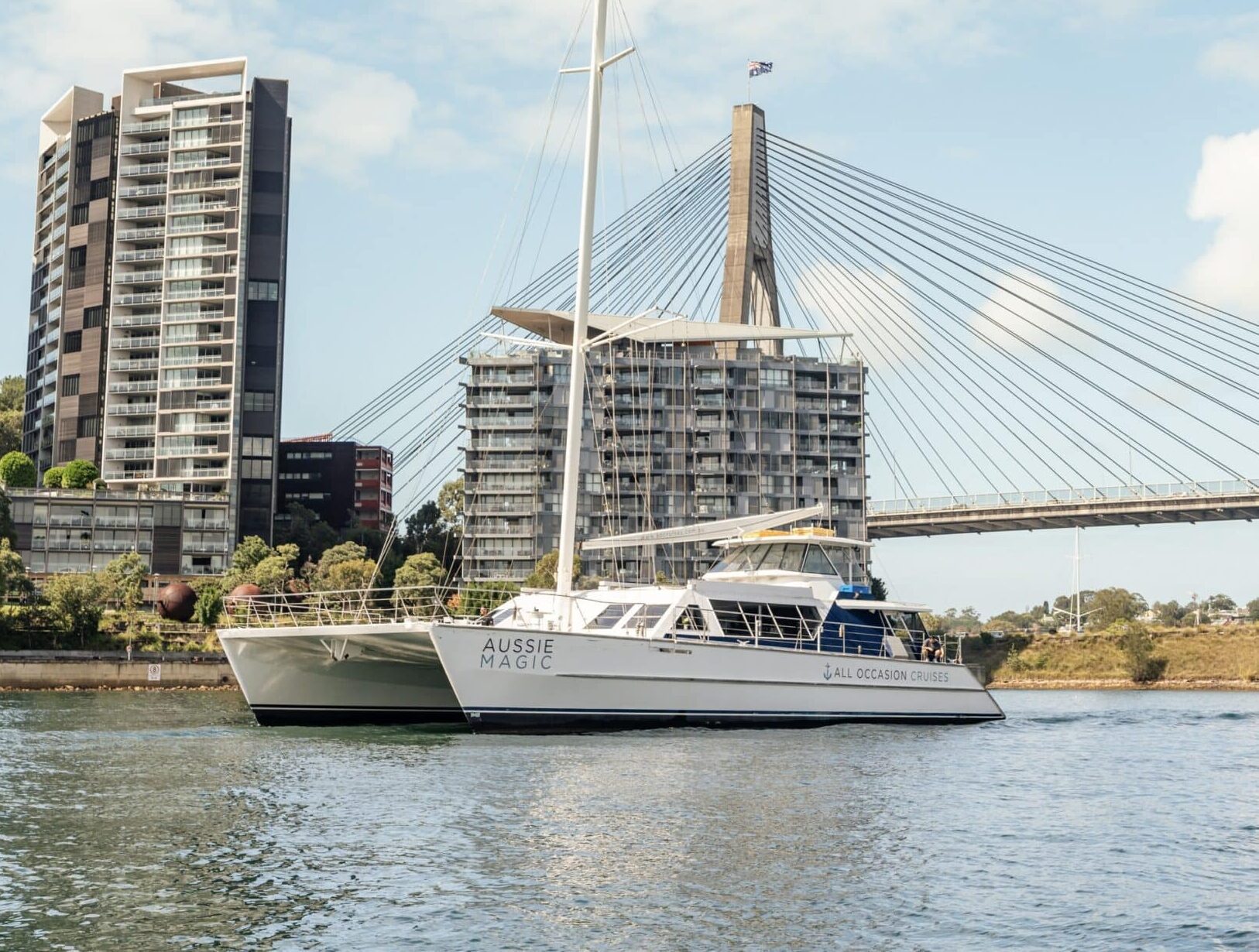 The white catamaran "Aussie Magic" sails past modern apartments and a cable-stayed bridge under a partly cloudy sky.