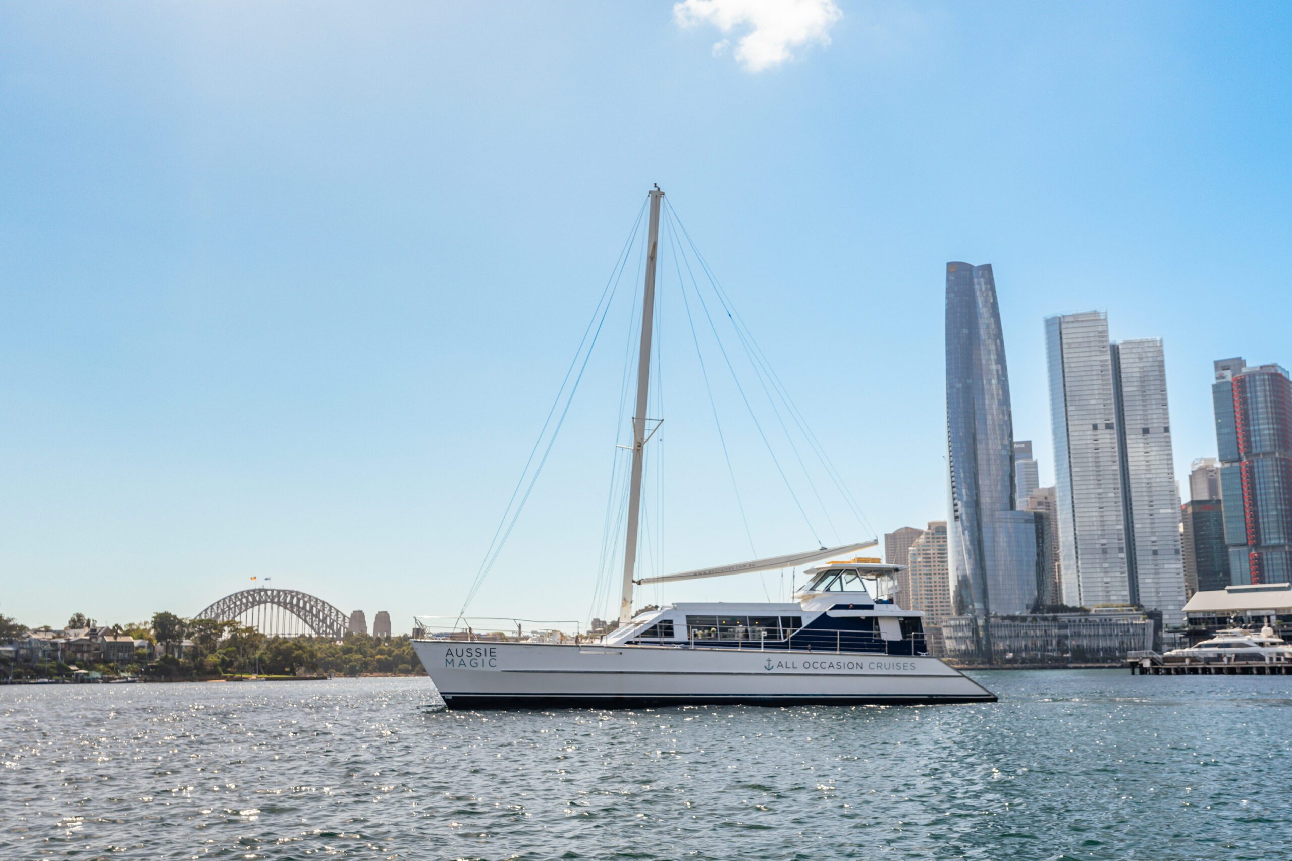 Aussie Magic, a sleek white catamaran, cruises through Sydney Harbour with the Sydney Harbour Bridge and city skyscrapers in the background beneath a clear blue sky.
